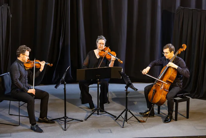 Drei Musiker spielen Violine und Cello auf einer Bühne vor Notenständern. Das Konzert 