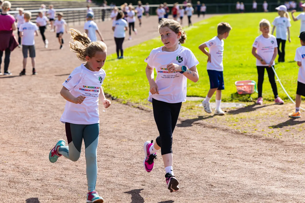 Zwei Mädchen laufen beim Spendenlauf der Bismarckschule Kierspe nebeneinander auf einer roten Tartanbahn. Sie tragen T-Shirts mit dem Aufdruck „SPONSORENLAUF Bismarckschule Kierspe“.