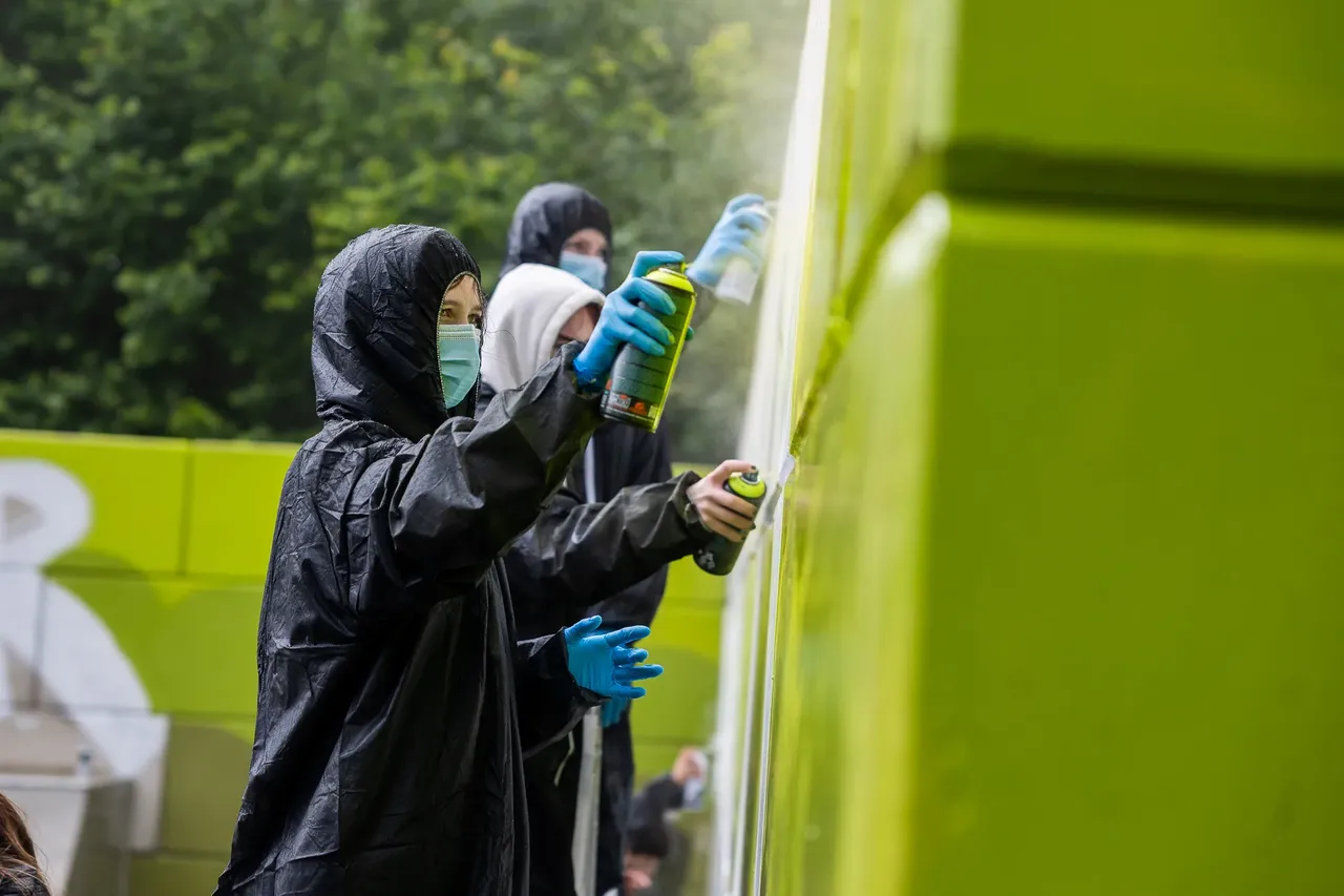 Jugendliche sprühen Graffiti auf eine grüne Wand an einer Skateranlage. Eine Person trägt eine dunkle Regenjacke mit Kapuze, eine Maske und blaue Handschuhe.