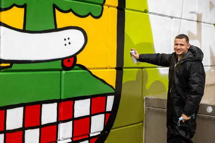 Ein lächelnder junger Mann mit dunkler Jacke sprüht ein farbenfrohes Graffiti mit einem Skateboard-Motiv auf eine Betonwand. Im Hintergrund sind weitere Graffiti-Elemente in Gelb, Grün und Rot zu sehen.