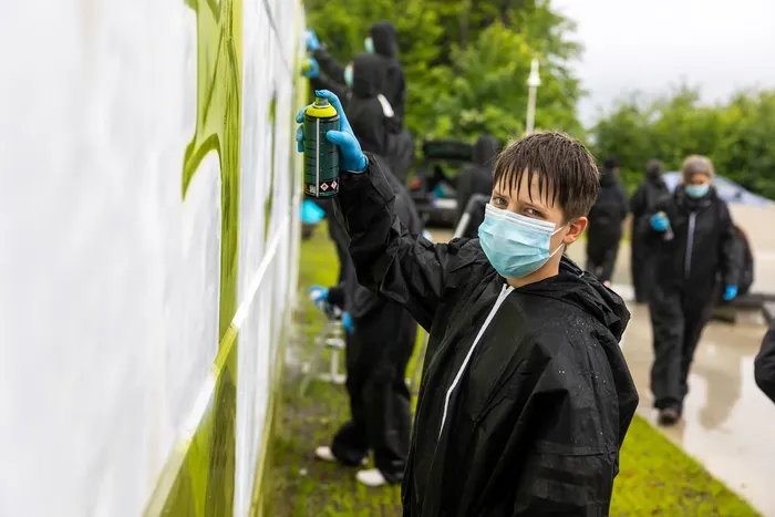 Ein Jugendlicher mit Schutzmaske sprüht mit einer Spraydose eine weiße Wand an. Im Hintergrund sind weitere Personen bei der Gestaltung einer Skateranlage zu sehen.