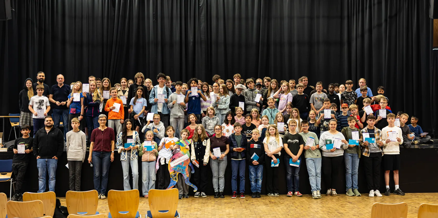 Eine Gruppe von Schülern und Lehrern steht auf einer Bühne vor einem dunklen Vorhang. Die Schüler halten Urkunden oder Auszeichnungen in den Händen.