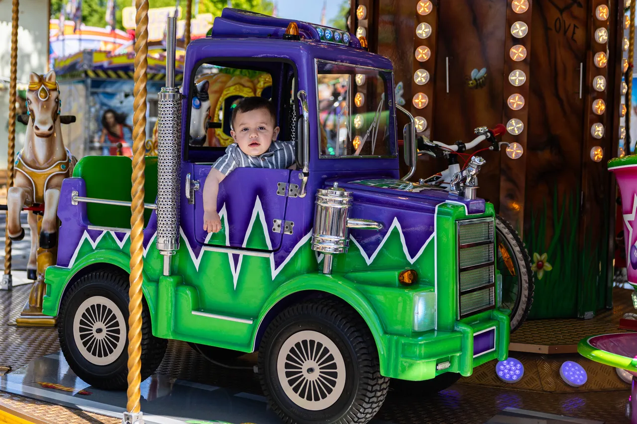 Kleiner Junge sitzt lächelnd in einem grün-violetten Karussellfahrzeug in Form eines Lastwagens.
