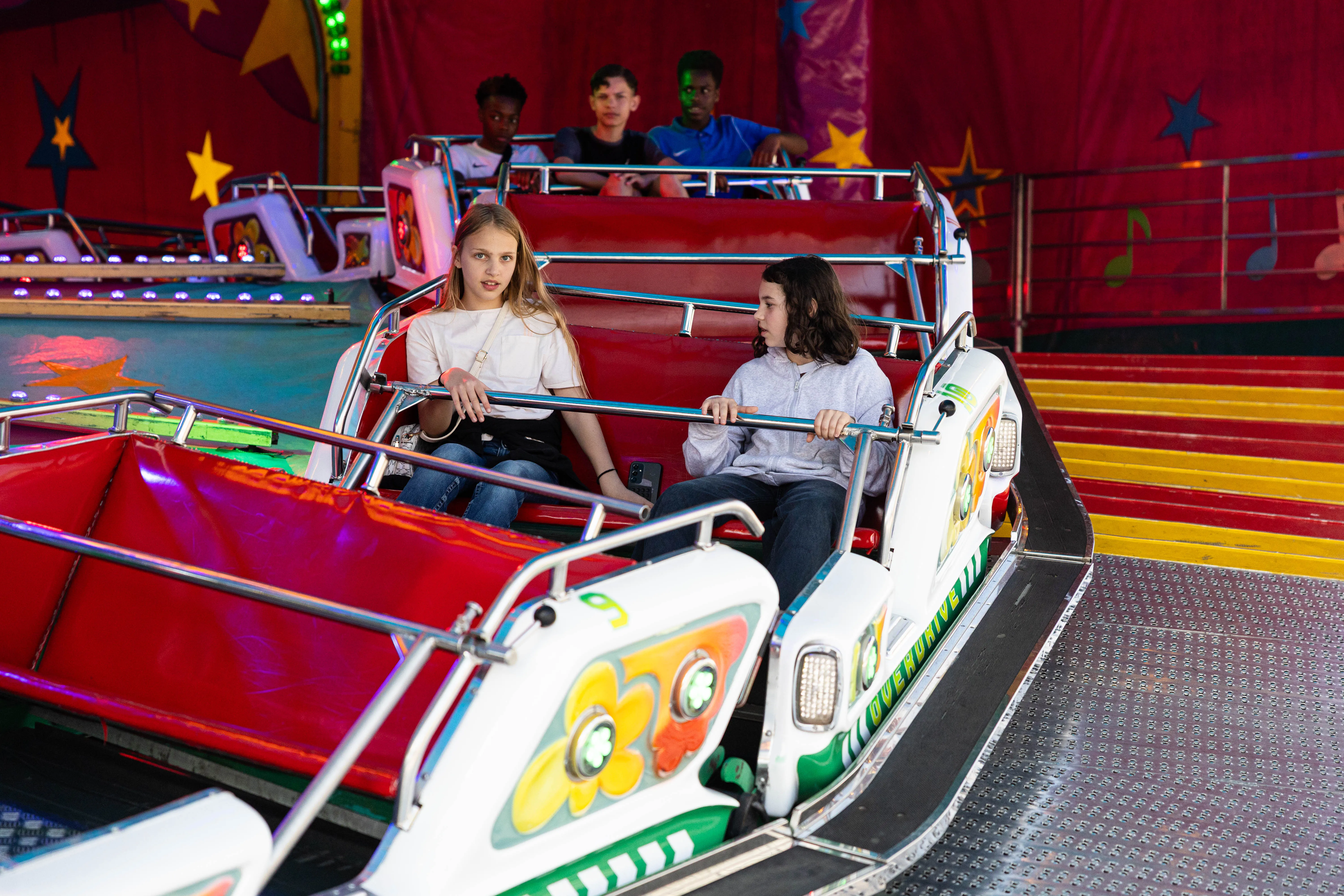 Zwei Kinder sitzen in einem Autoscooter auf der Kirmes, während im Hintergrund weitere Besucher zu sehen sind.