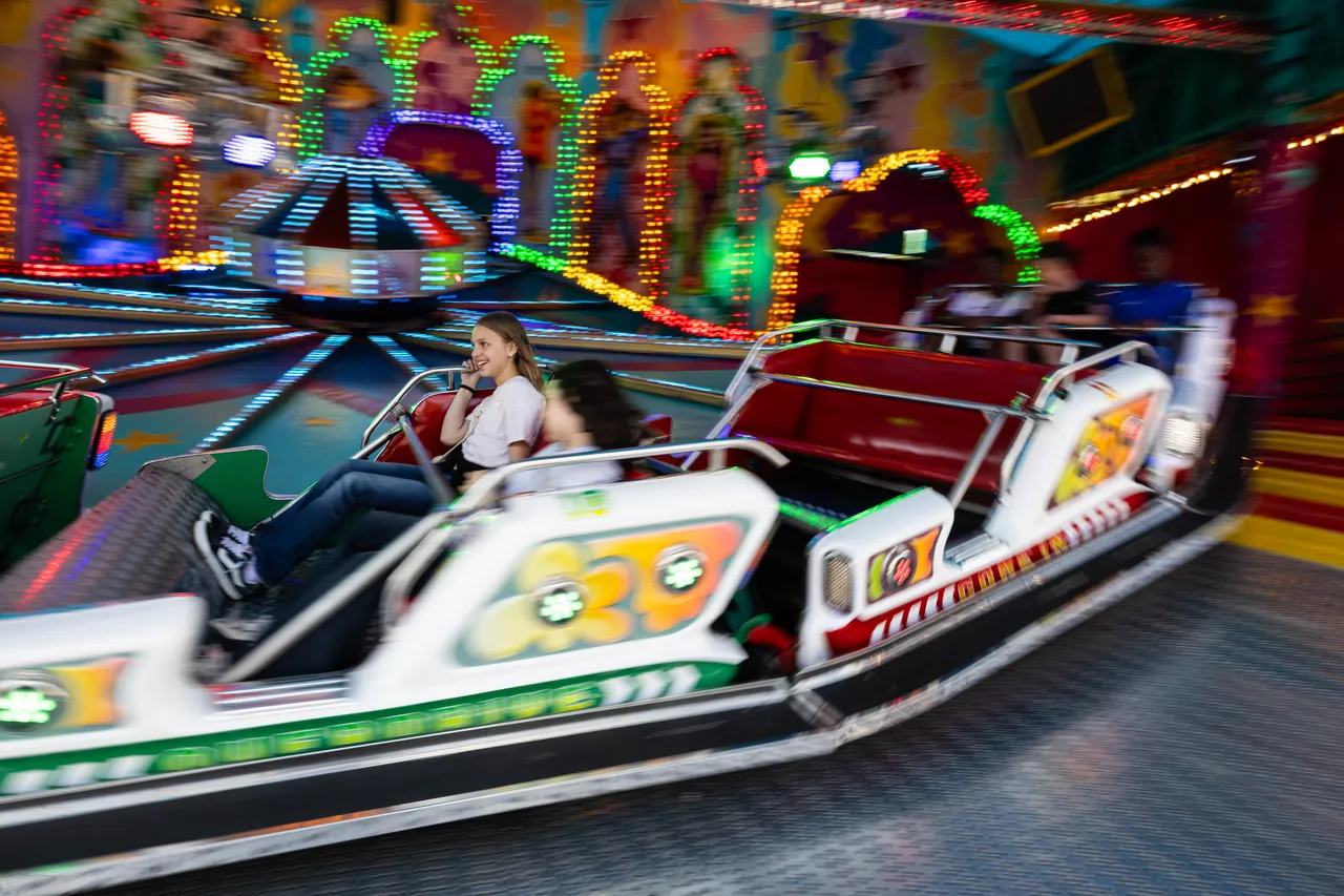 Fahrende Menschen in einem Karussell auf der Kirmes, im Hintergrund bunte Lichter und weitere Fahrgeschäfte.
