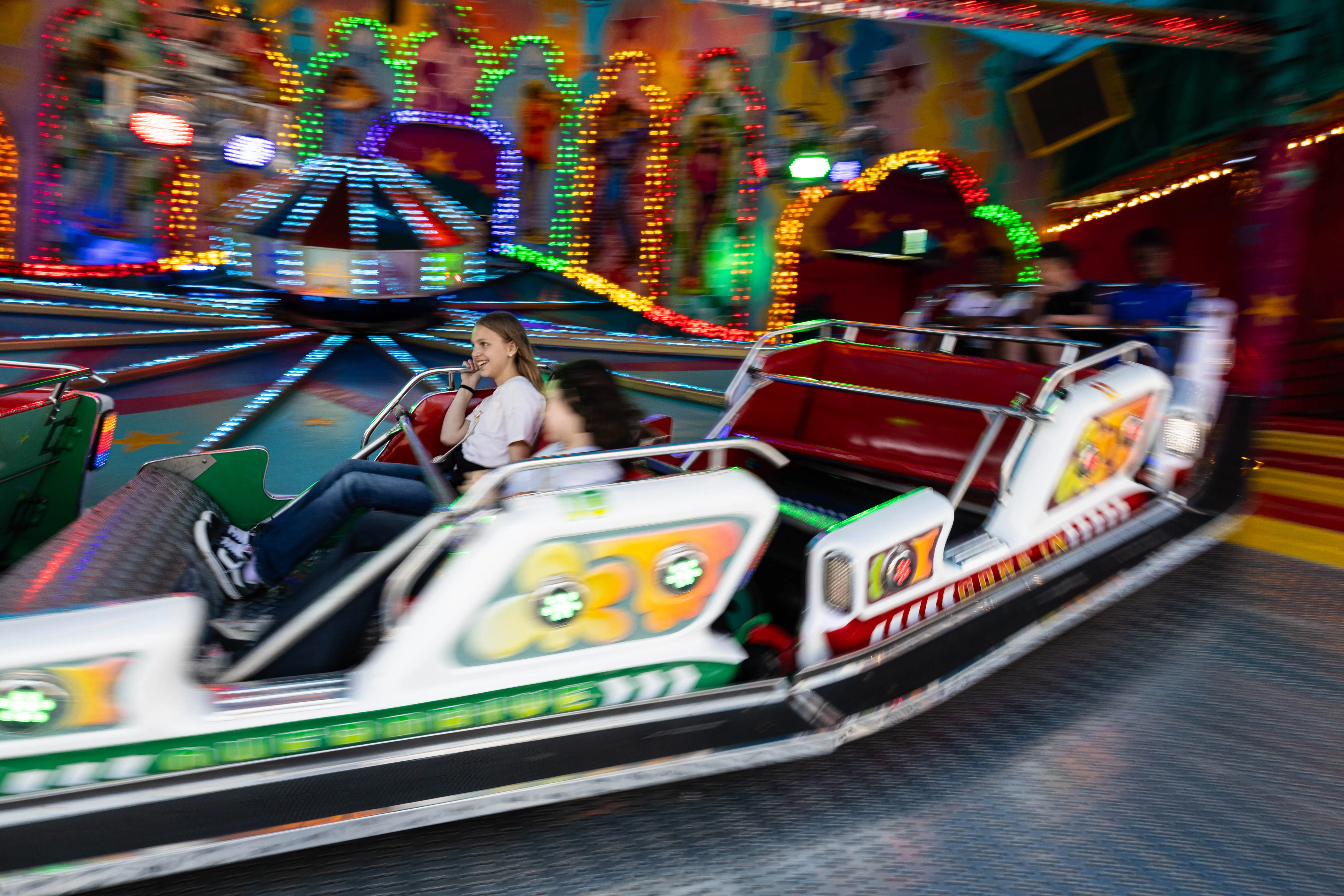 Fahrende Menschen in einem Karussell auf der Kirmes, im Hintergrund bunte Lichter und weitere Fahrgeschäfte.