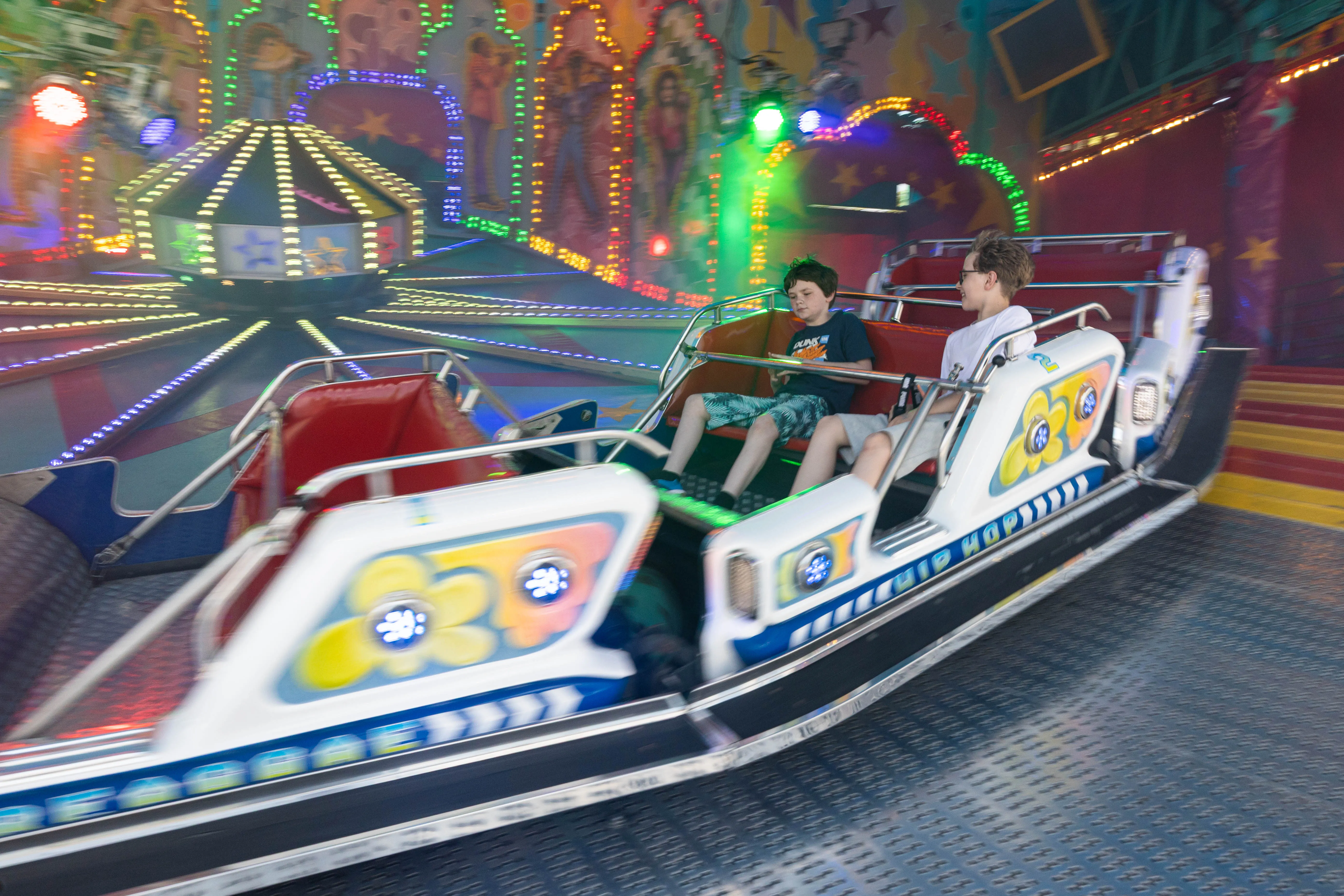 Zwei Jungen sitzen in einem Fahrgeschäft auf der Kirmes, während es sich dreht. Im Hintergrund sind weitere Lichter und Attraktionen zu sehen.