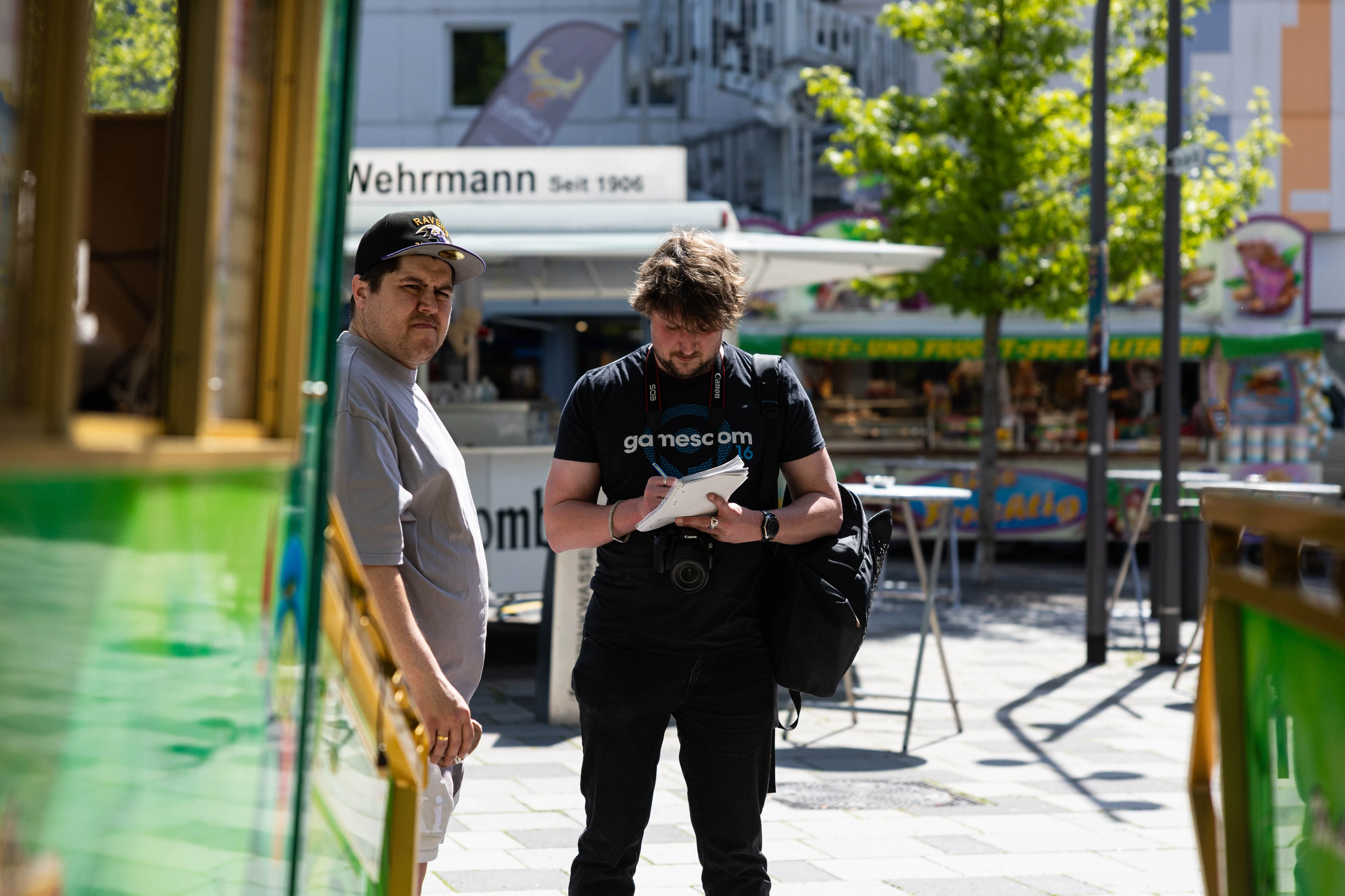 Zwei Männer betrachten ein Smartphone in der Fußgängerzone während der Meinerzhagener Pfingskirmes. Im Hintergrund sind Geschäfte und Cafés zu sehen.