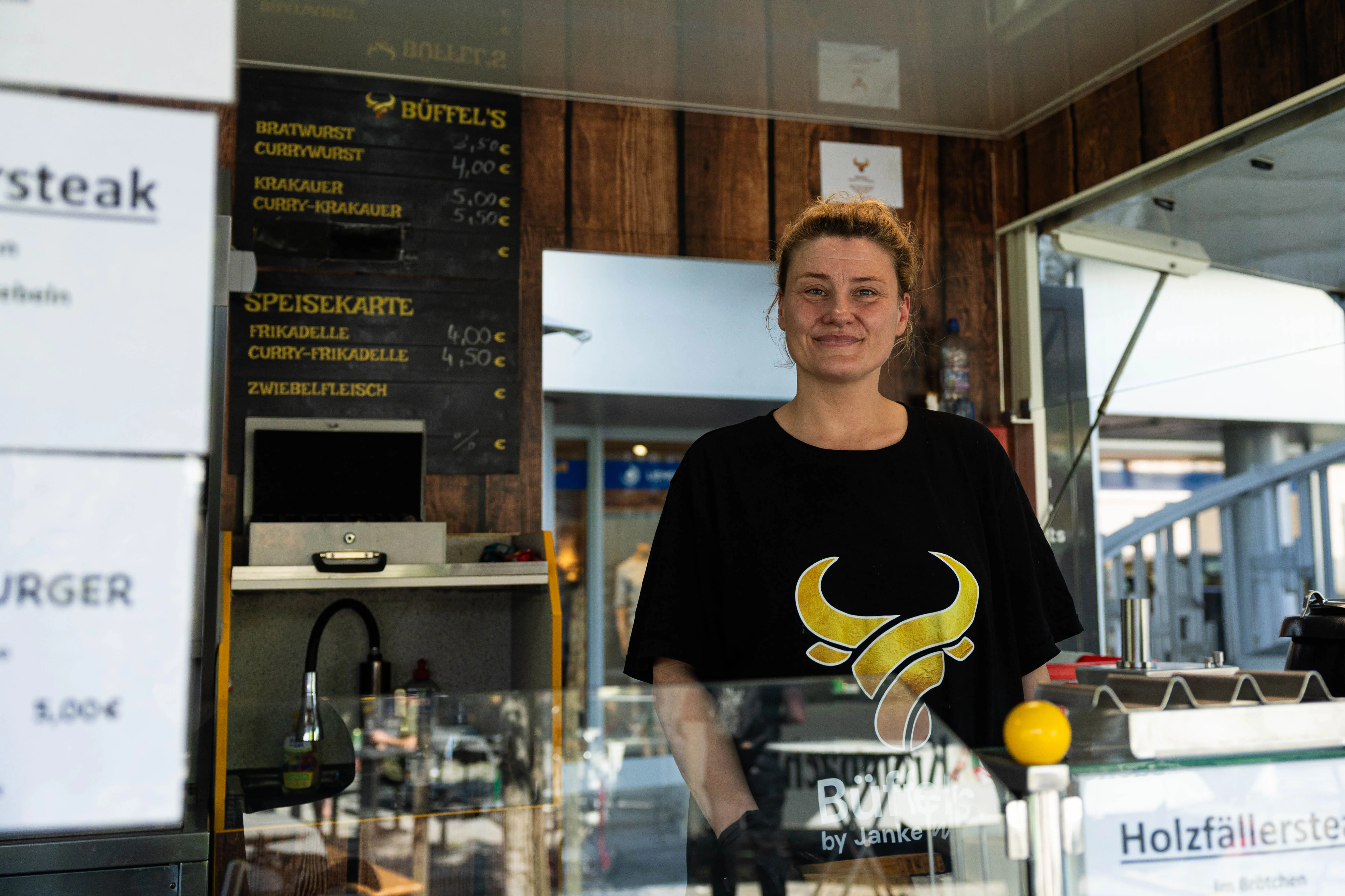 Junge Frau lächelt hinter dem Verkaufstresen eines Imbissstandes mit der Aufschrift „Büffel’s“ und einer Speisekarte mit Preisen für Bratwurst und Currywurst.