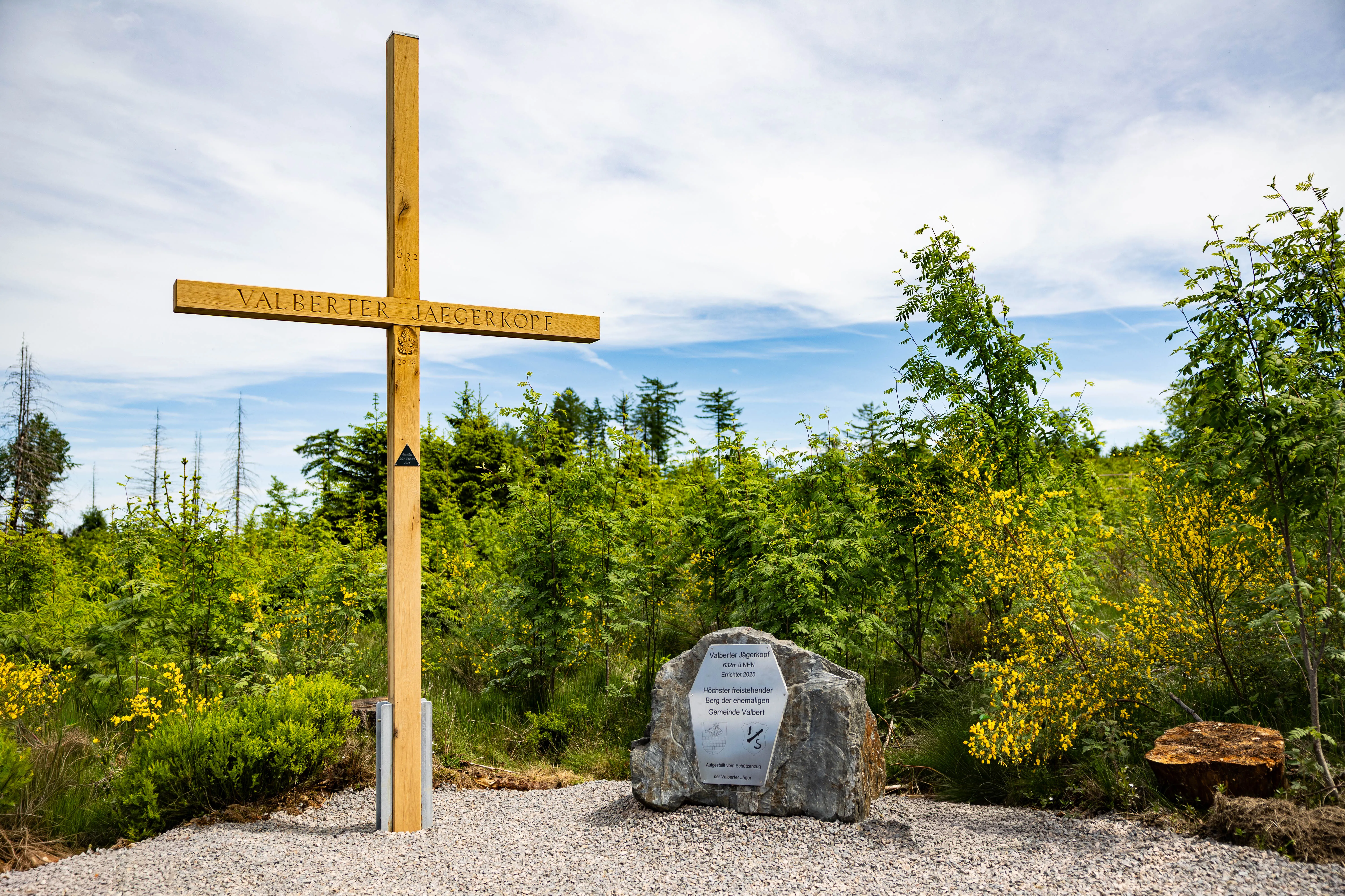 Holzkreuz mit dem Namen „Valberter Jägerkopf“ vor einem Felsblock mit Gedenktext und blühenden Büschen unter blauem Himmel.