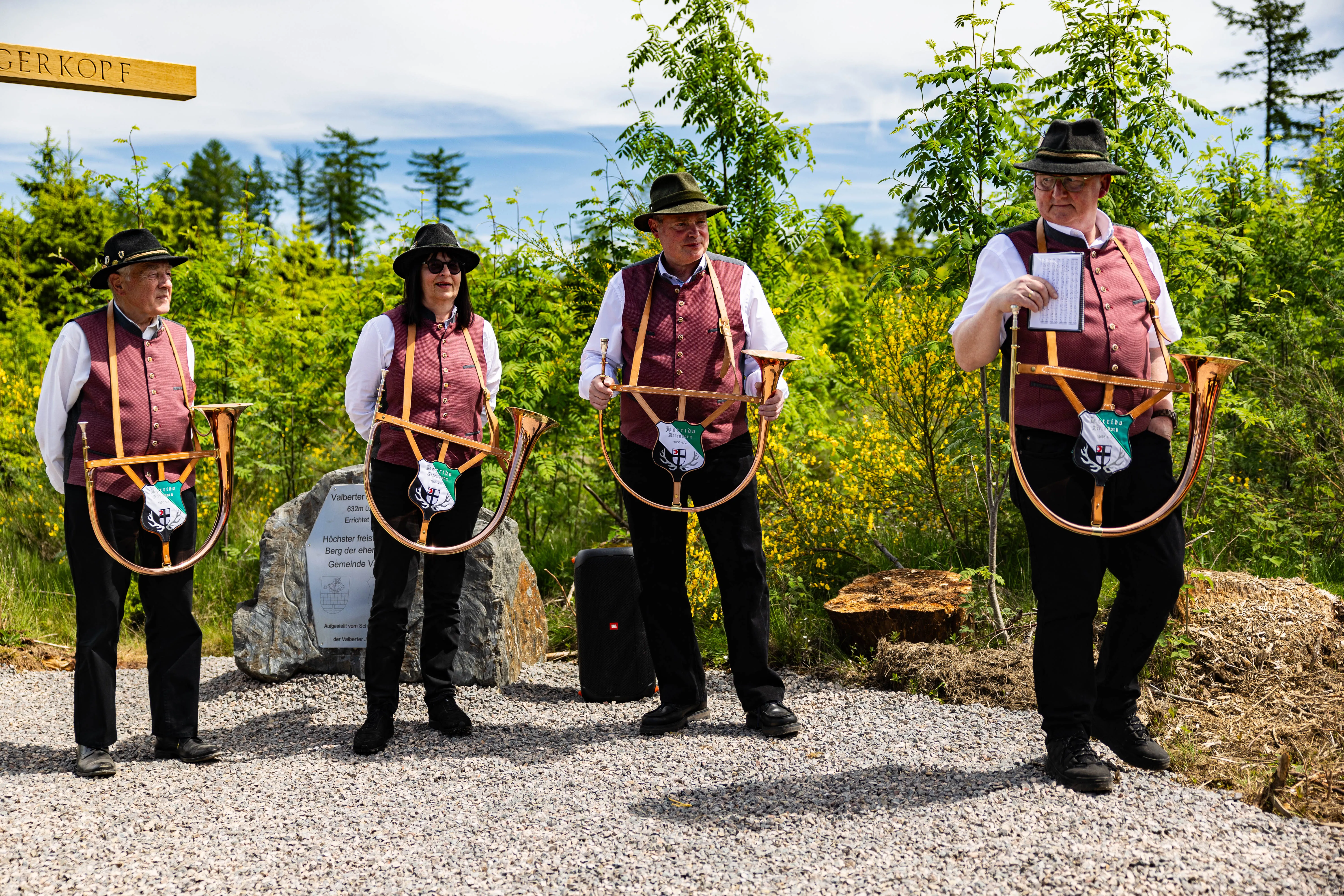 Vier Personen in traditioneller Tracht spielen auf großen Alphörnern vor einer Waldkulisse und einem Wegweiser zum Valberter Jägerkopf.