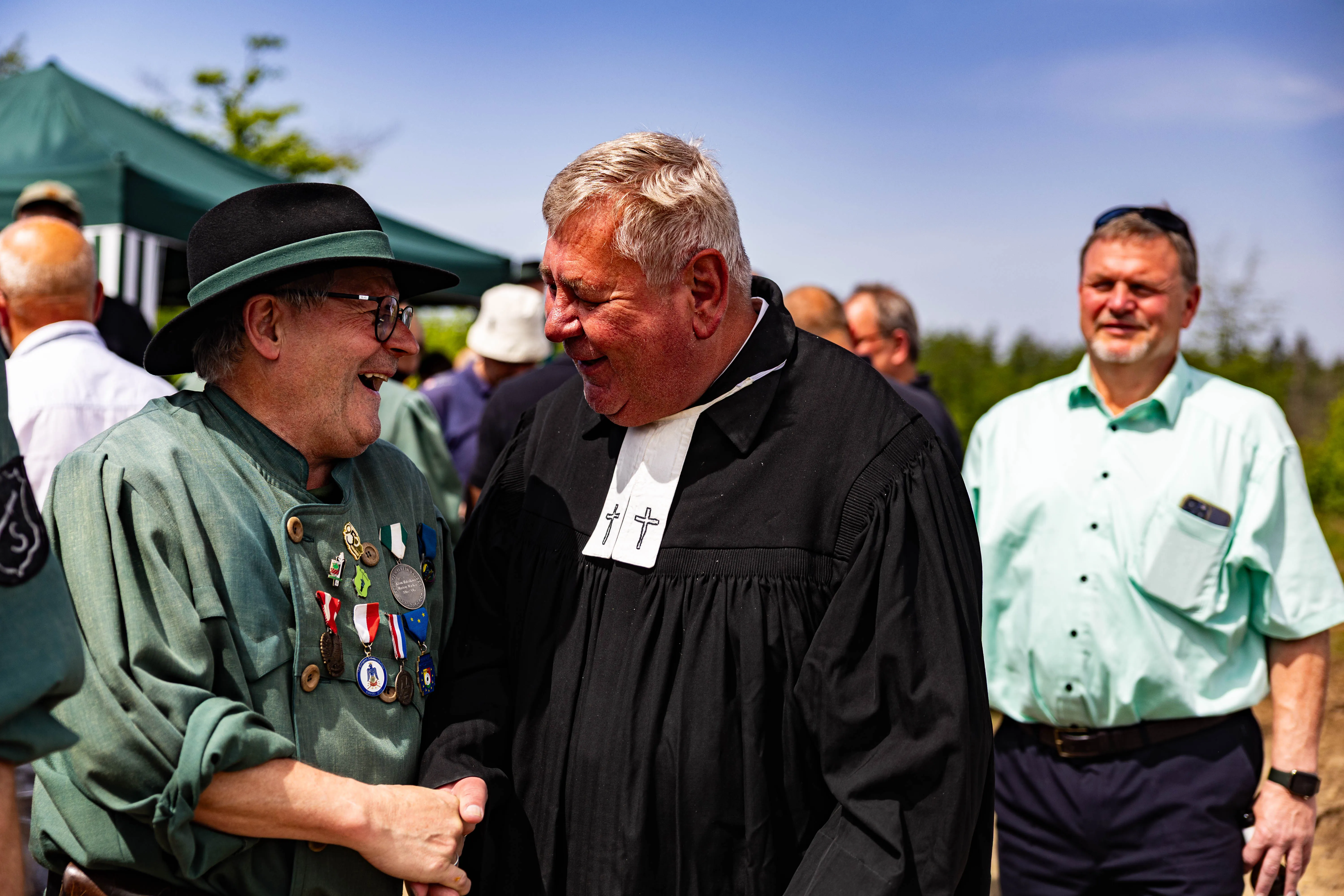 Zwei lächelnde Männer, einer in Uniform mit Orden, der andere in schwarzer Soutane mit weißem Kragen, begrüßen sich. Im Hintergrund weitere Personen und Zelte.