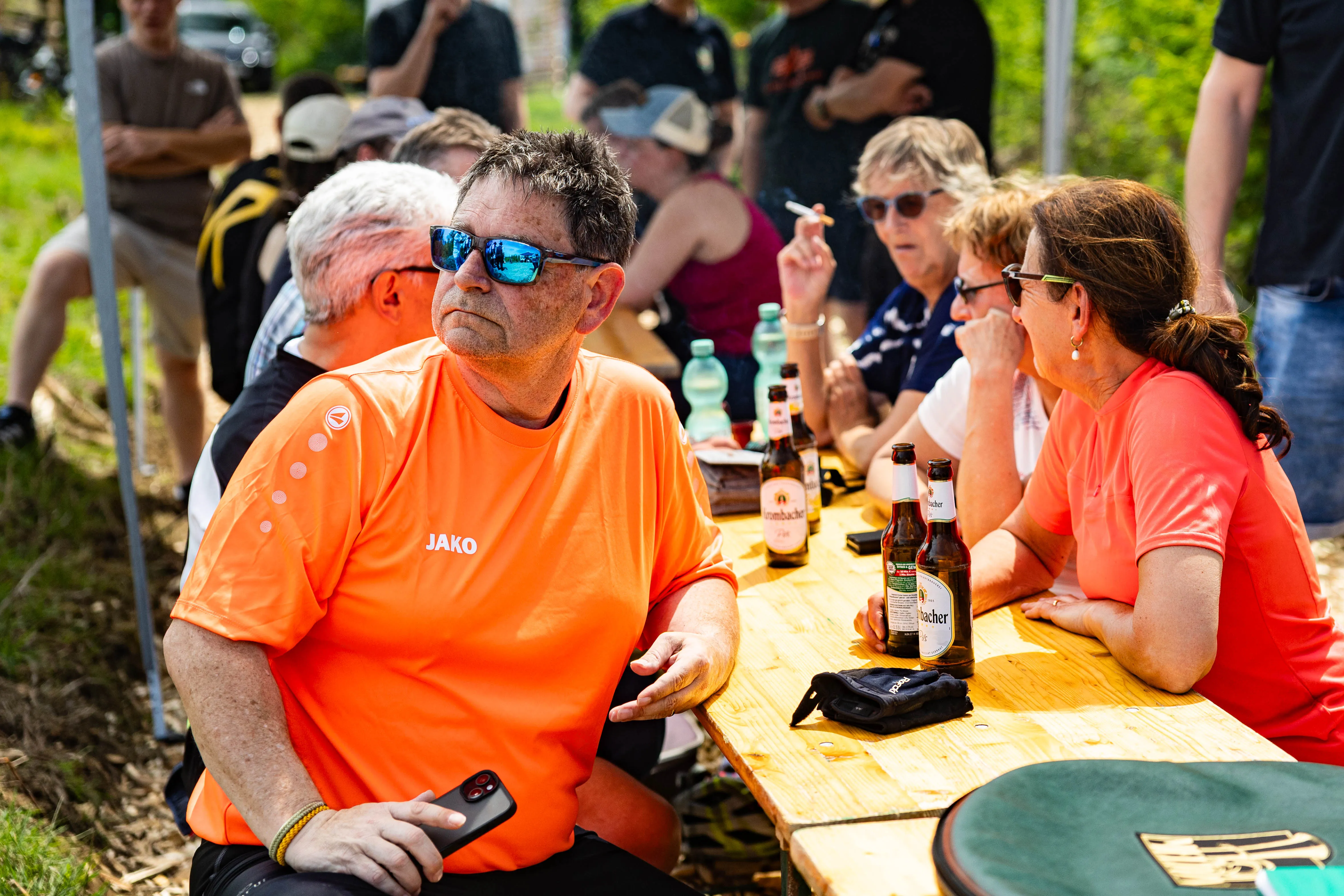 Ein Mann mit Sonnenbrille und orangefarbenem T-Shirt sitzt an einem Biertisch, während im Hintergrund weitere Personen bei einer Veranstaltung zusammensitzen und Getränke konsumieren.