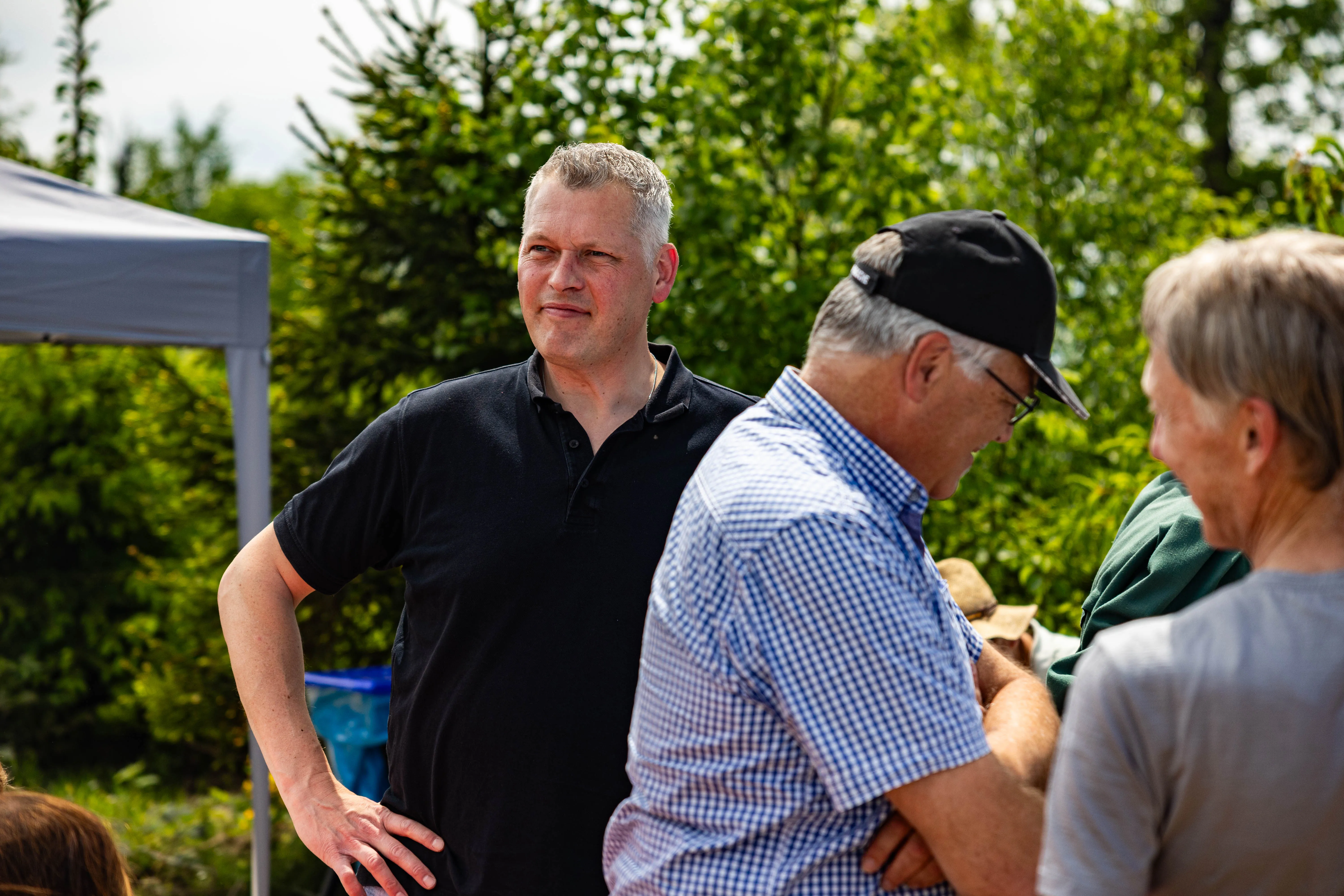 Ein Mann mit grau meliertem Haar blickt zur Seite, während er mit der Hand in die Hüfte gestützt steht. Im Hintergrund sind weitere Personen und ein Zelt zu sehen.