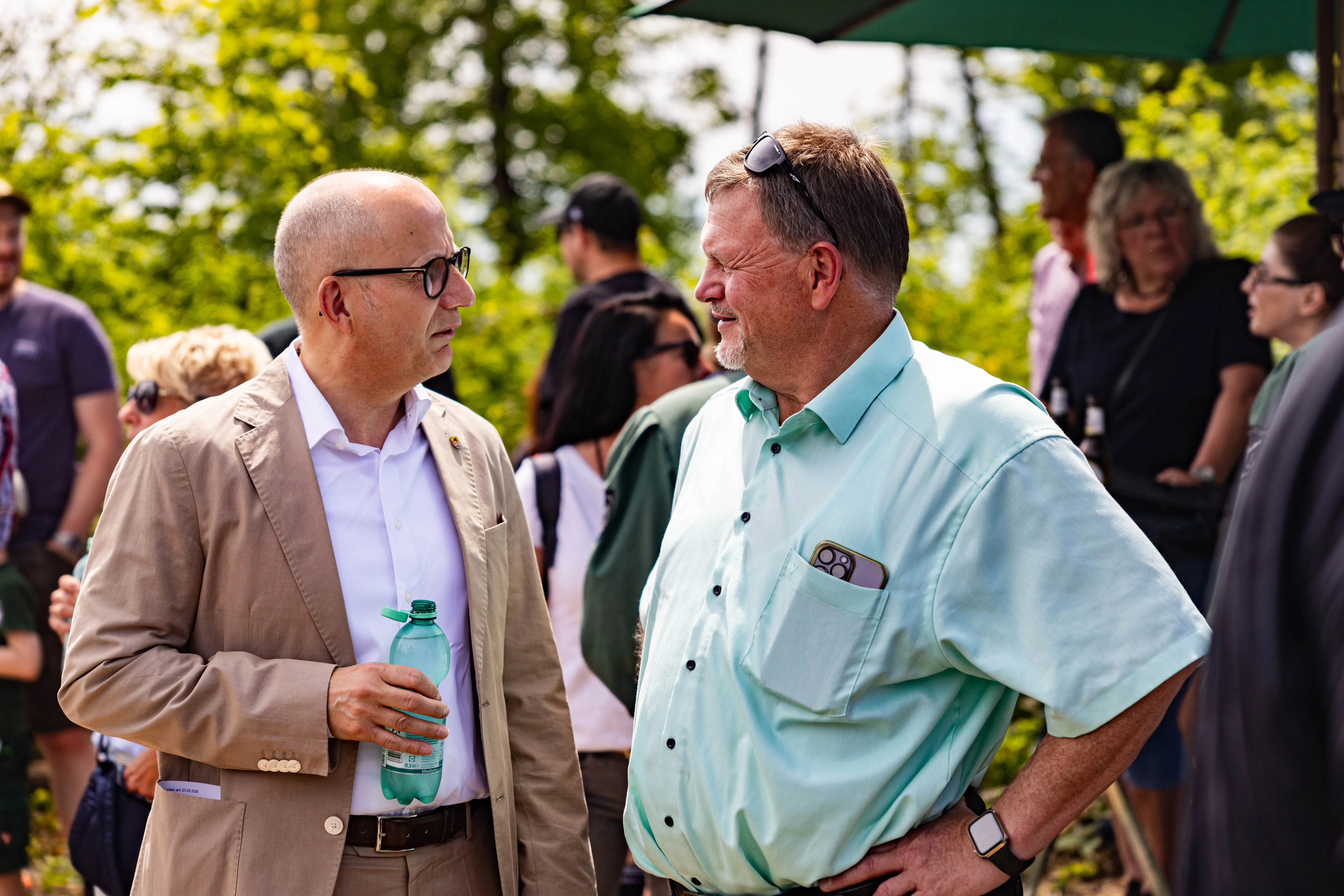 Zwei Männer im Gespräch während einer Freiluftveranstaltung, im Hintergrund weitere Menschen und grüne Bäume.