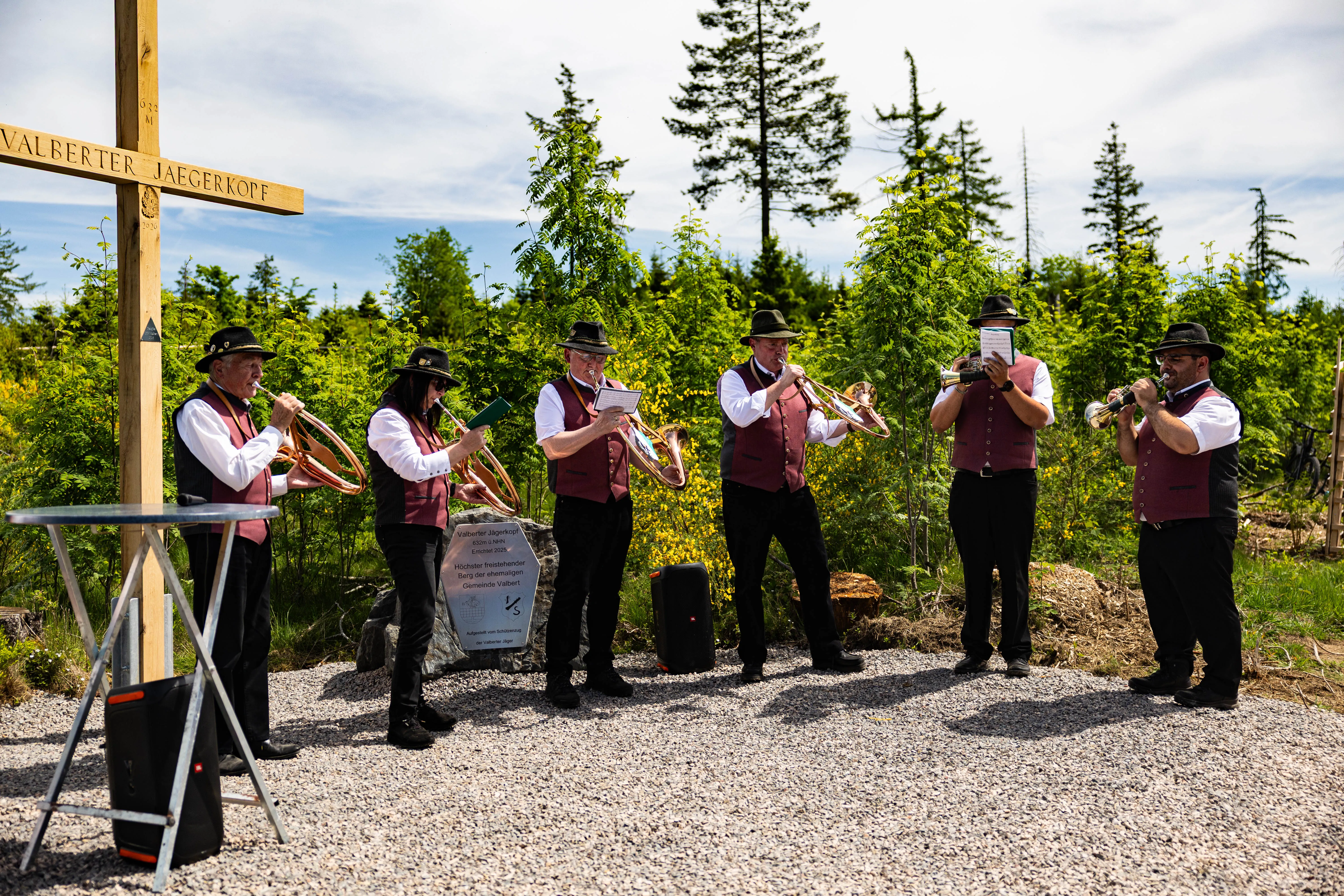 Blaskapelle spielt vor einem Holzkreuz im Wald. Ein Schild mit Text steht zwischen den Musikern.