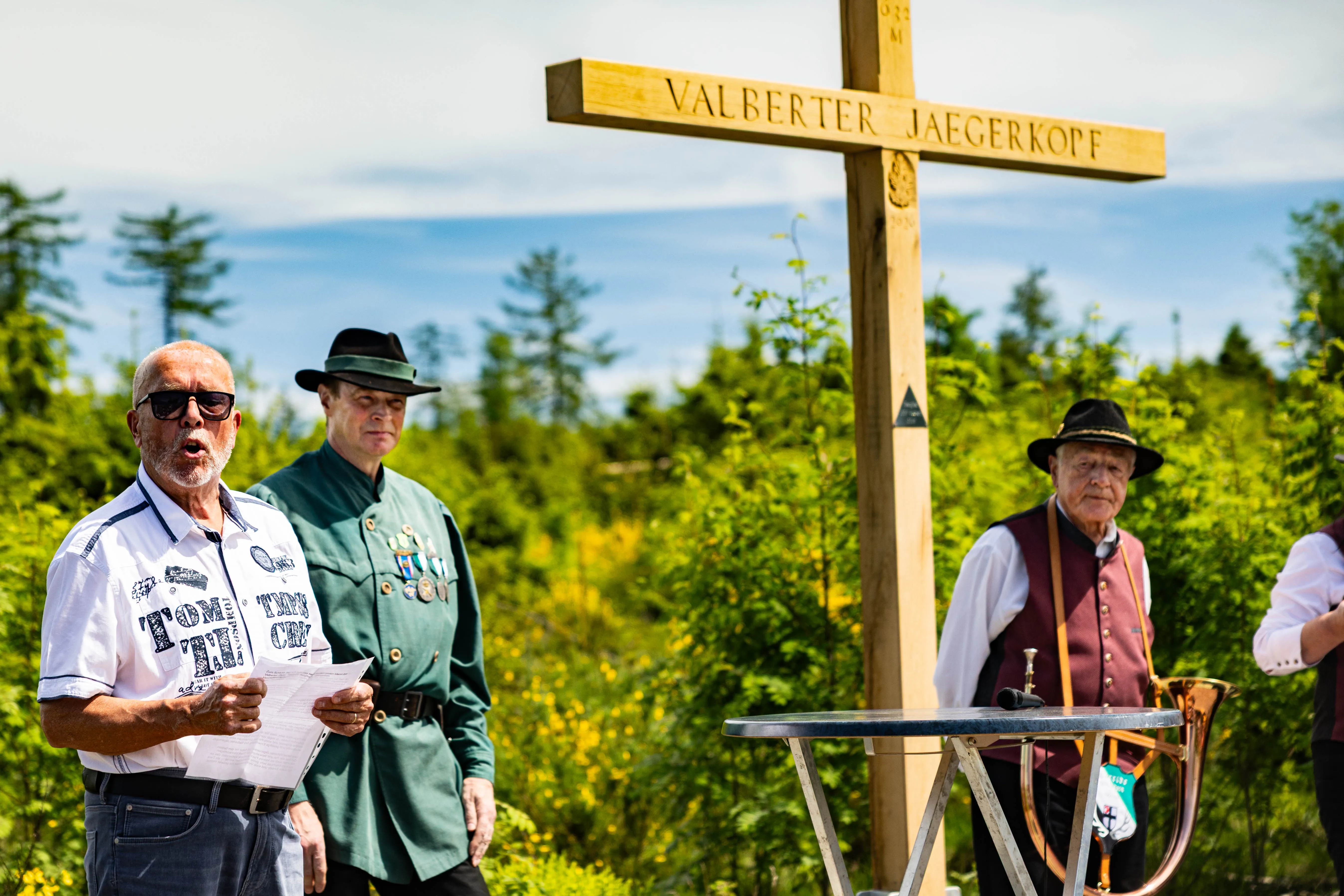 Ein Mann hält während eines Gottesdienstes ein Blatt Papier und spricht in ein Mikrofon, während zwei weitere Männer im Hintergrund stehen. Im Hintergrund ein Holzkreuz mit der Aufschrift „Valberter Jägerkopf“.