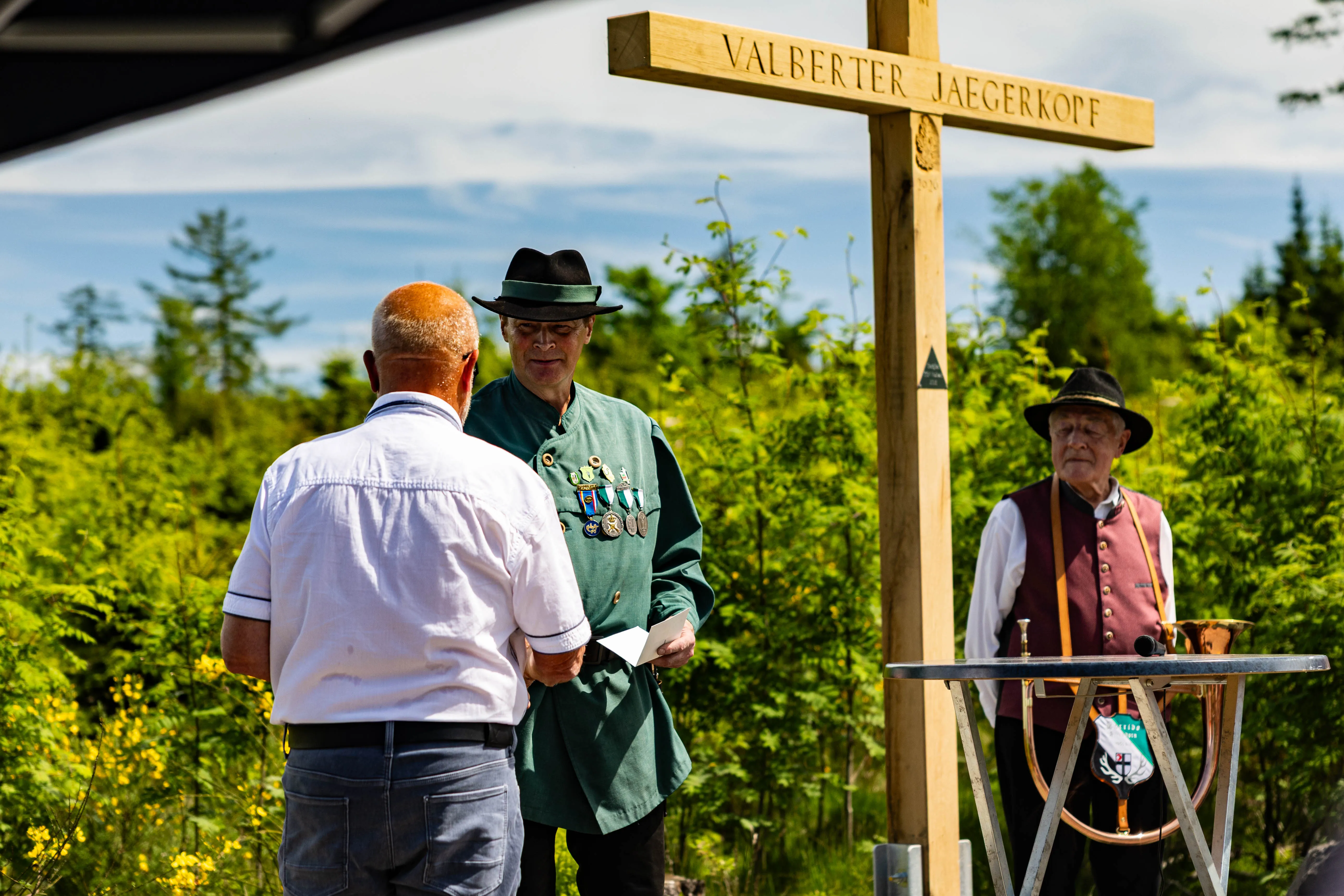 Zwei Männer in traditioneller Tracht stehen vor einem Holzkreuz mit der Inschrift „Valberter Jägerkopf“, während ein dritter Mann im Hintergrund eine Trommel spielt.