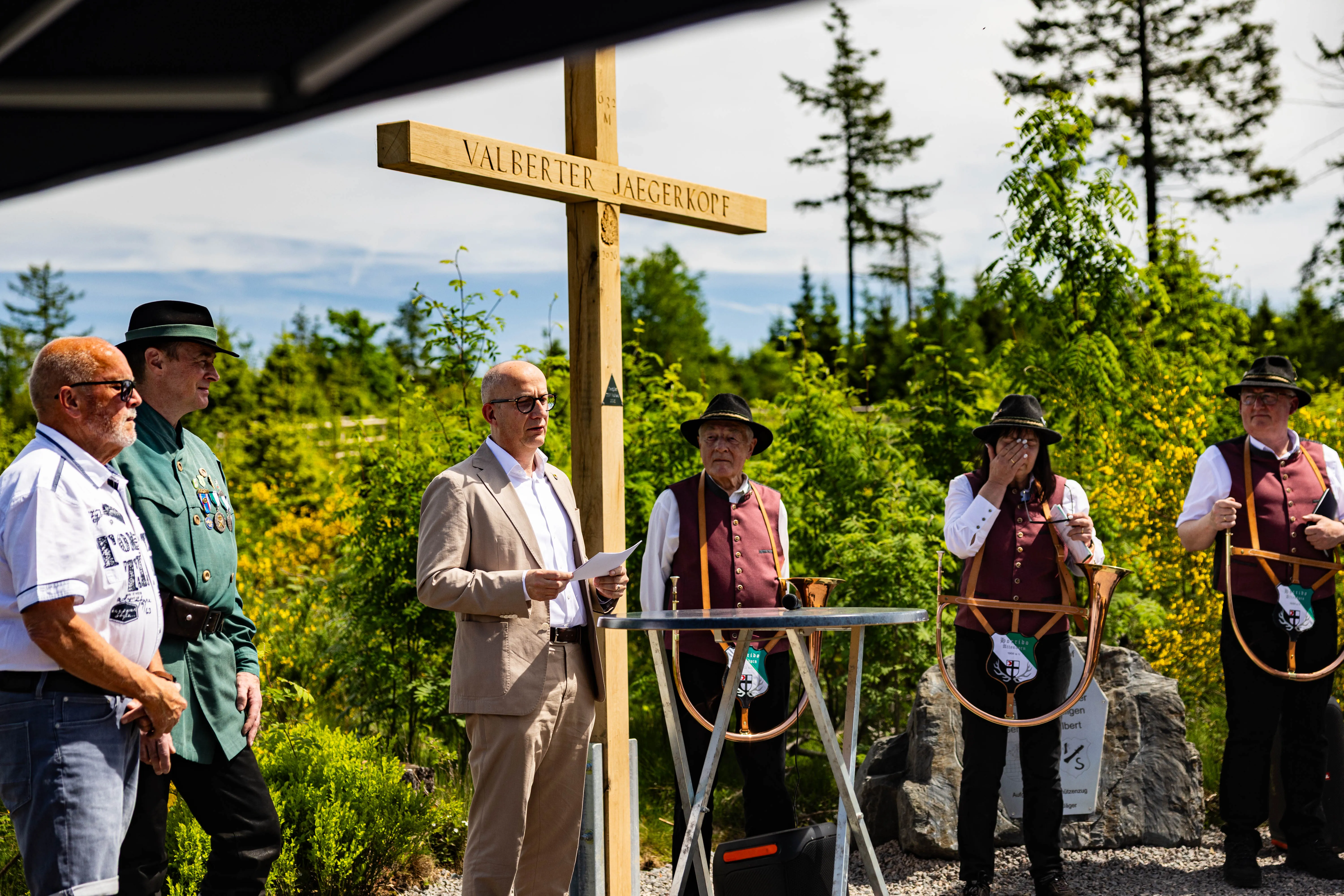 Mehrere Männer in traditioneller Tracht und Anzug stehen vor einem Holzkreuz mit der Aufschrift „Valberter Jägerkopf“ in einer Waldlandschaft.