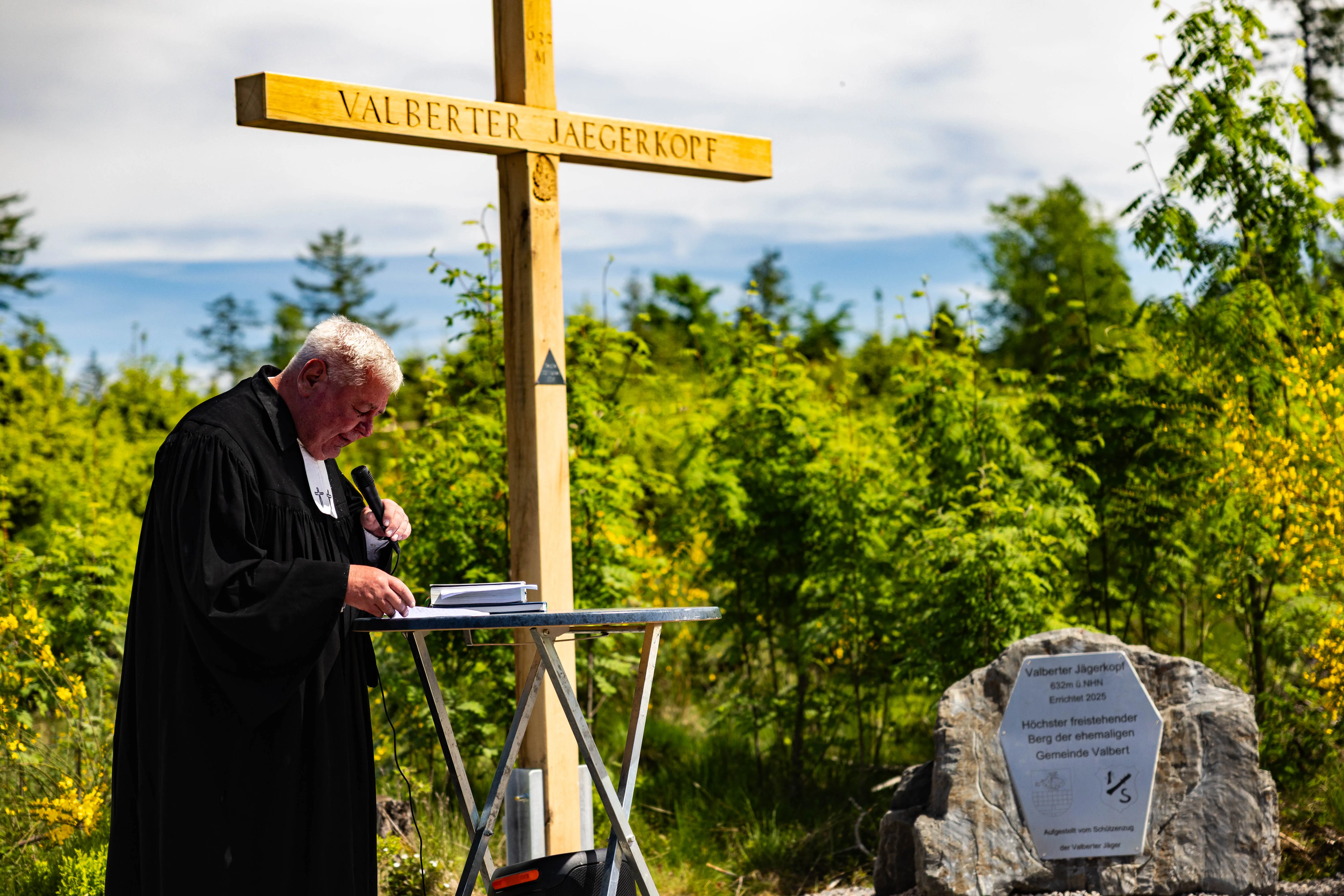 Ein Priester in schwarzer Robe hält während eines Eröffnungsgottesdienstes am Valberter Jägerkopf eine Ansprache vor einem Holzkreuz. Im Hintergrund eine gelb blühende Landschaft und ein Gedenkstein.