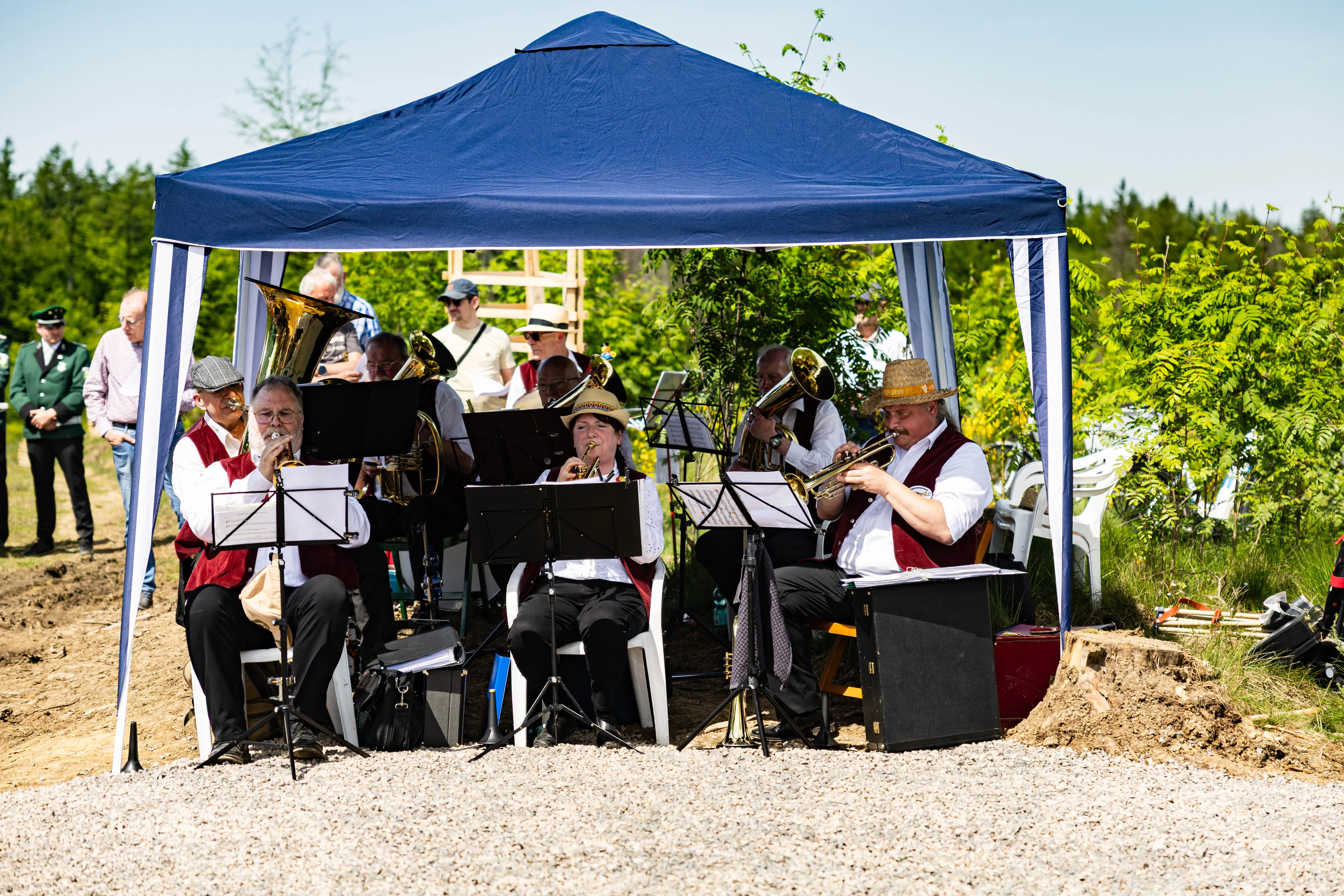Blasmusikensemble spielt unter einem blauen Pavillon im Freien, während im Hintergrund Zuschauer stehen. Im Hintergrund sind Bäume und ein Waldstück zu sehen.