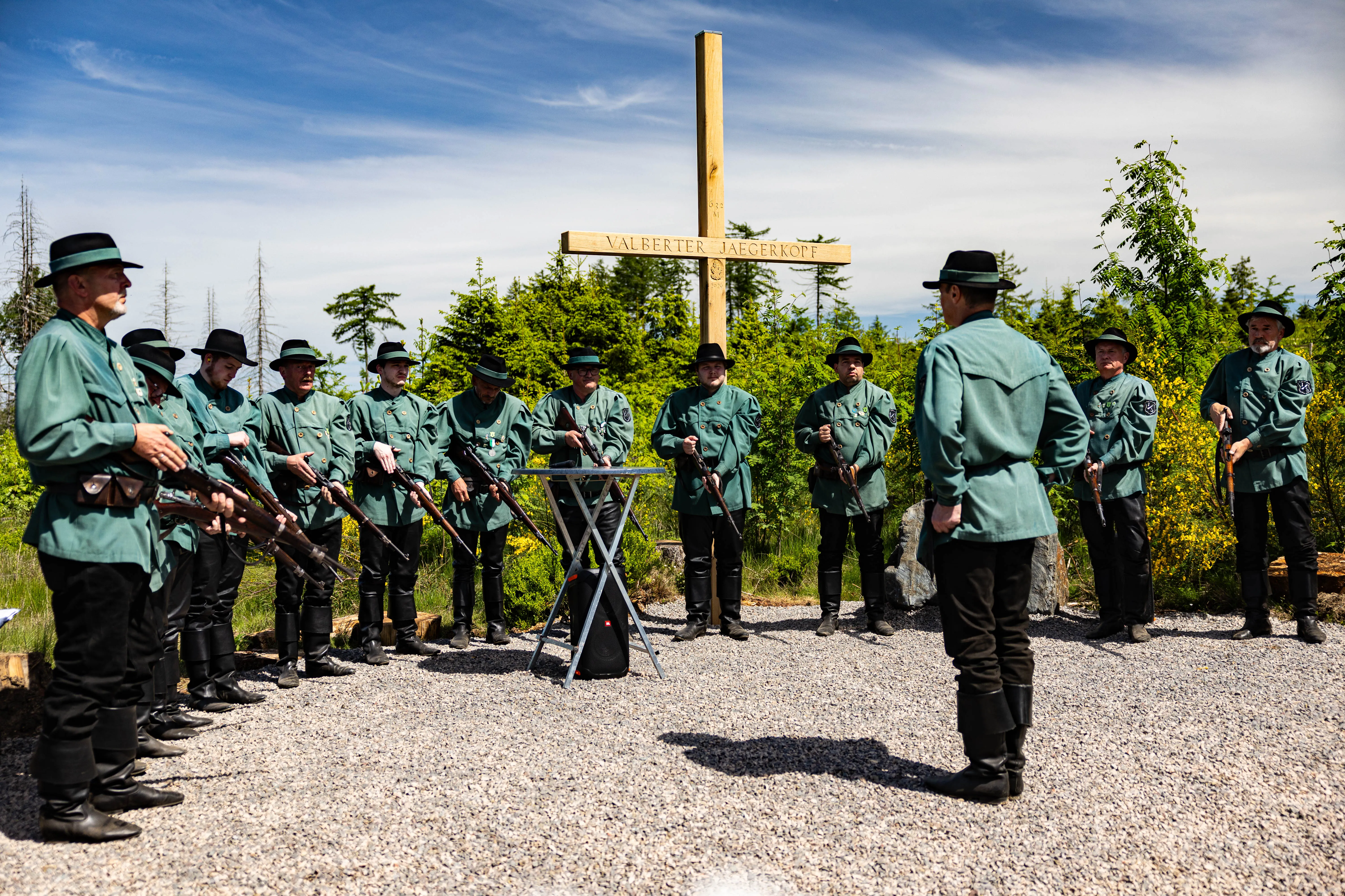 Eine Gruppe von Männern in traditionellen Jägeruniformen mit Gewehren steht um ein Podest mit Notenblättern vor einem Holzkreuz auf einem Kiesplatz. Im Hintergrund Bäume und ein blauer Himmel.