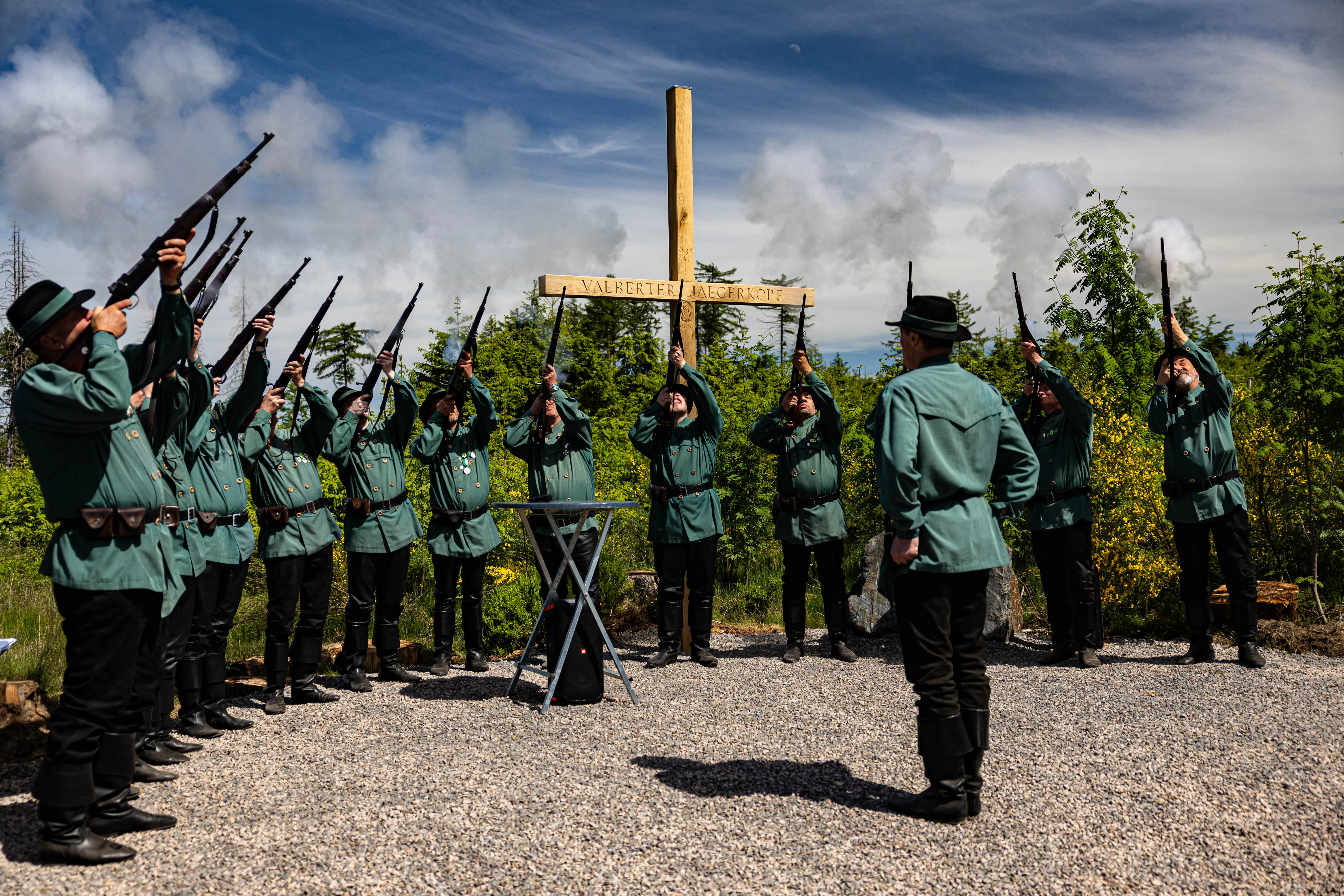 Eine Gruppe von Männern in historischen Uniformen mit Gewehren bei einer Zeremonie vor einem hölzernen Kreuz mit der Inschrift „Valberter Jägerkopf“.