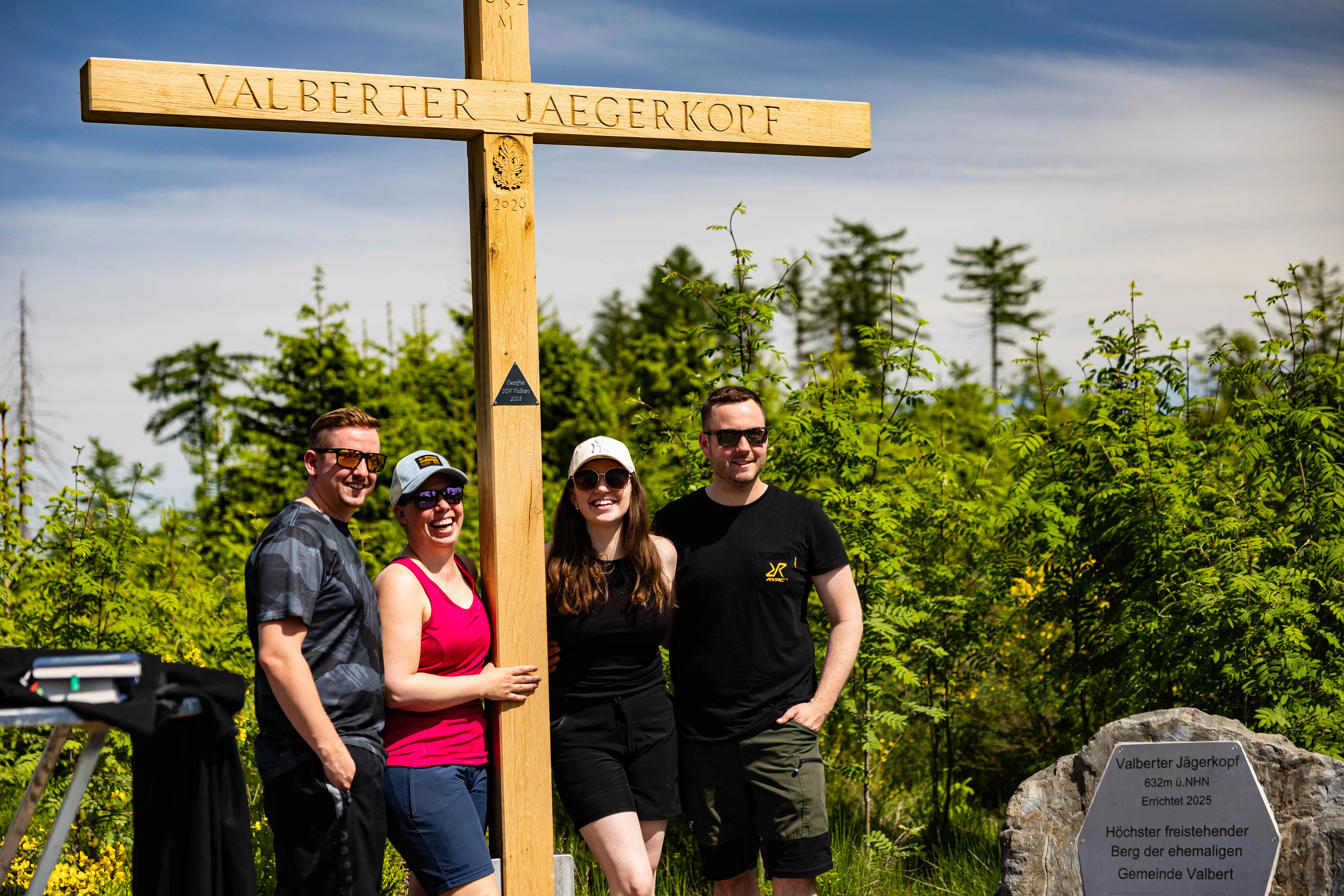 Vier junge Erwachsene stehen vor einem großen Holzkreuz auf dem Valberter Jägerkopf, im Hintergrund ein dichter Wald und ein blauer Himmel.