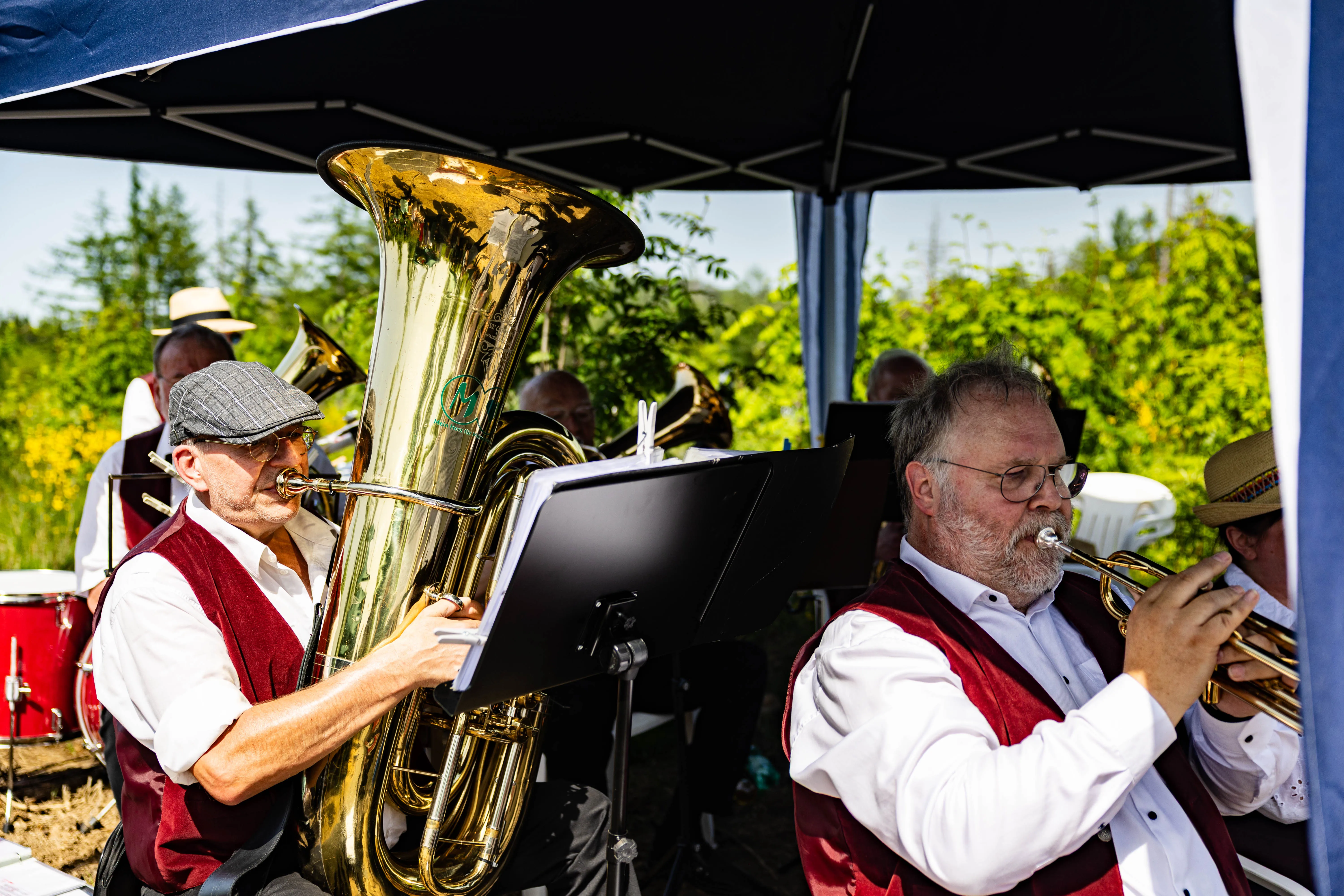 Zwei Musiker spielen während eines Eröffnungsgottesdienstes Posaune und Tuba unter einem Pavillon. Im Hintergrund weitere Musiker und grüne Vegetation.