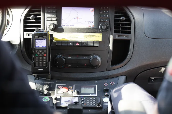Cockpit eines Polizeifahrzeugs mit verschiedenen Funkgeräten und einem Navigationssystem, das eine Karte zeigt.