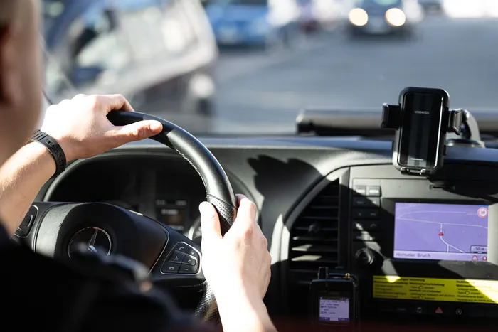 Person hält Lenkrad eines Autos fest, während ein Navigationsgerät und ein Smartphone am Armaturenbrett befestigt sind. Im Hintergrund ist eine Straße mit parkenden Autos zu sehen.
