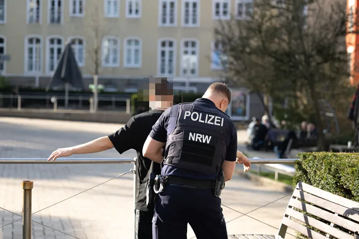 Ein Polizist der Polizei NRW kontrolliert eine Person an einem Geländer in einer Fußgängerzone.