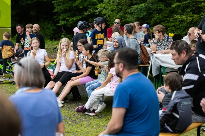 Zuschauer sitzen und stehen auf einer Rasenfläche vor einer Gruppe von Menschen, darunter Kinder mit Eis. Im Hintergrund sind Bäume und ein Wald zu sehen.