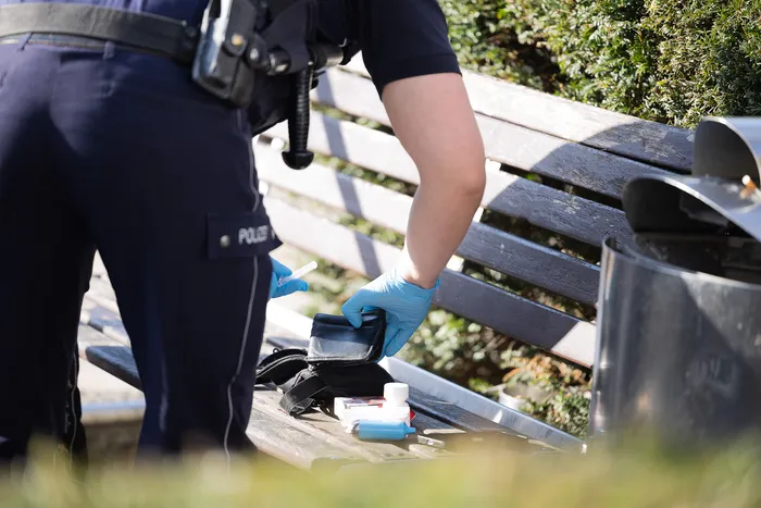 Ein Polizist mit blauen Handschuhen sammelt auf einer Parkbank Spritzen und andere Utensilien in einer schwarzen Tasche.