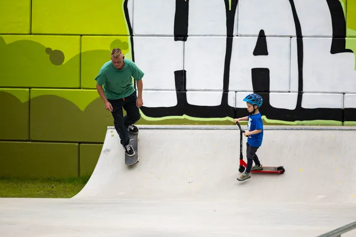 Ein Mann fährt mit einem Skateboard eine Rampe im neu eröffneten Skatepark in Halver hinunter, während ein Junge mit einem Tretroller auf ihn zukommt. Im Hintergrund eine graue Wand mit Graffiti und eine grüne Wand.