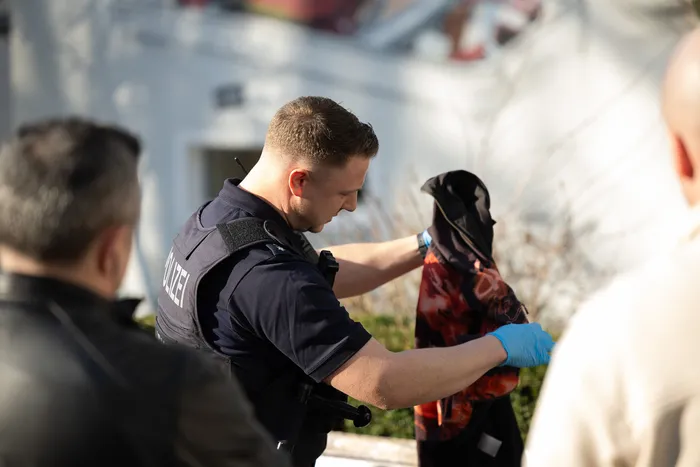 Ein Polizist in Uniform und mit blauen Handschuhen untersucht den Inhalt einer schwarzen Tasche im Freien, während ein Kollege im Hintergrund steht.