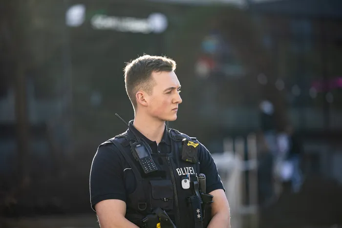 Ein Polizist in Uniform mit Funkgerät und Ausrüstung blickt nachdenklich in die Ferne, im Hintergrund verschwommene Menschen und Fahrzeuge.