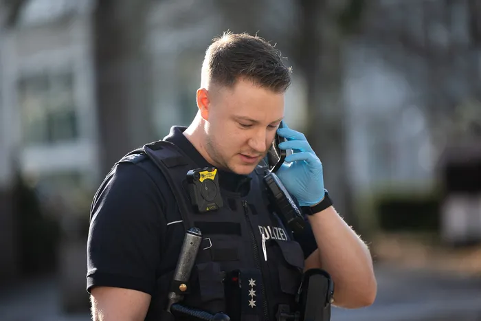 Polizist mit Schutzweste und Handschuhen telefoniert ernst. Er trägt ein Bodycam-Gerät auf der Brust.