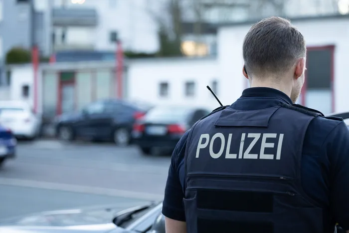 Polizist mit der Aufschrift „POLIZEI“ auf der Schutzweste steht einer Straße mit parkenden Autos zu.