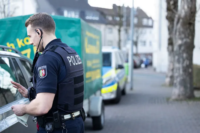 Ein Polizist mit Schutzweste und Headset spricht mit dem Fahrer eines silbernen Autos, während im Hintergrund weitere Polizeifahrzeuge stehen.