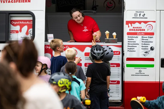 Eine Eisverkäuferin steht lächelnd an einem Eiswagen und reicht zwei jungen Mädchen Eiswaffeln über den Tresen. Im Hintergrund stehen weitere Kinder und warten. Der Eiswagen ist mit Preisen und Sorten dekoriert.