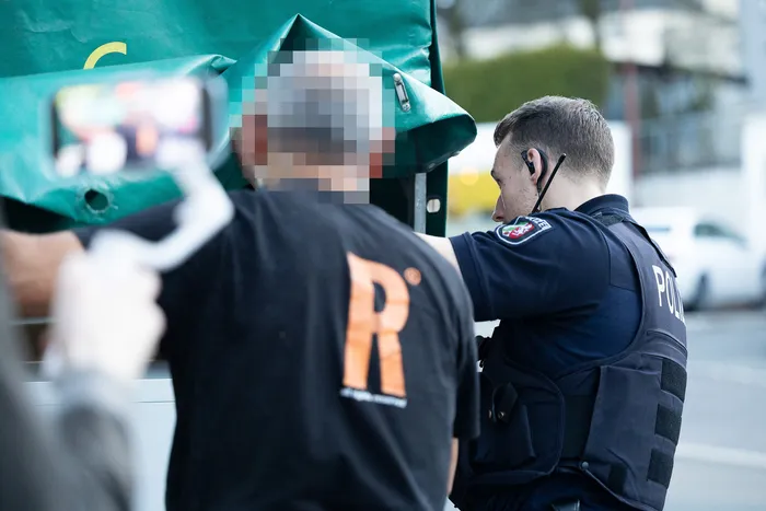 Polizist mit Schutzweste kontrolliert Person an einer Absperrung, deren Gesicht verpixelt ist. Im Hintergrund sind weitere Personen und Fahrzeuge zu sehen.