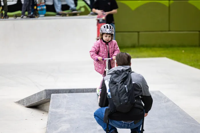 Ein kleines Kind mit Helm fährt auf einem Tretroller durch den neu eröffneten Skatepark in Halver, während eine erwachsene Person hockend daneben steht und zusieht. Im Hintergrund sind weitere Skater und die Parkanlage zu sehen.