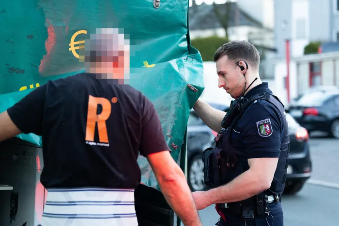 Ein Polizist kontrolliert eine Person vor einem Hintergrund mit einem Euro-Zeichen. Der Polizist trägt eine Uniform und ein Headset, die Person ein rotes T-Shirt mit einem weißen Buchstaben 'R'.