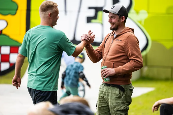 Zwei Männer geben sich vor einem Graffiti-Hintergrund die Hände. Im Hintergrund sind weitere Personen und Skateboards zu sehen.