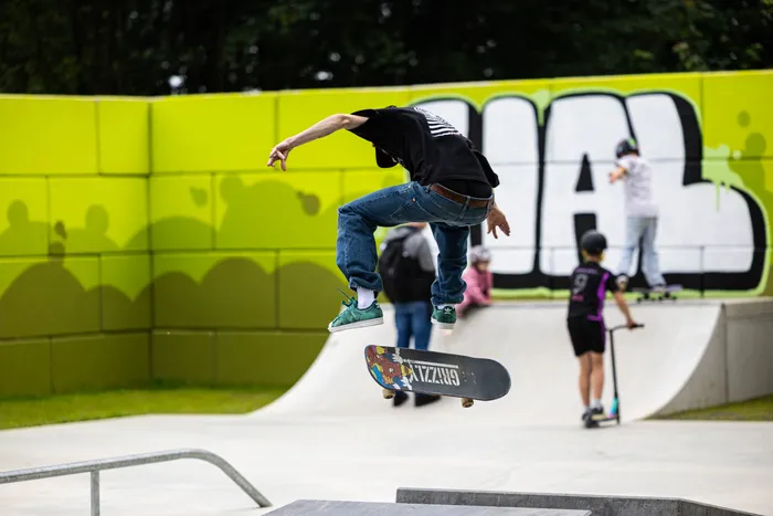 Ein Skater springt mit seinem Board in der Luft, während im Hintergrund andere Jugendliche auf Rollern und Skateboards fahren. Der Skatepark ist von einer grünen Mauer und Graffiti verziert.