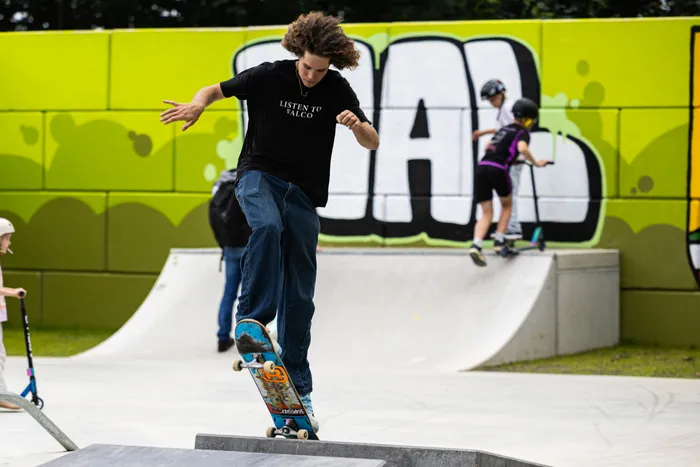 Ein junger Mann springt mit einem Skateboard über eine Rampe in einem Skatepark. Im Hintergrund ist ein weiteres Kind beim Skateboarden zu sehen, vor einer Wand mit Graffiti.