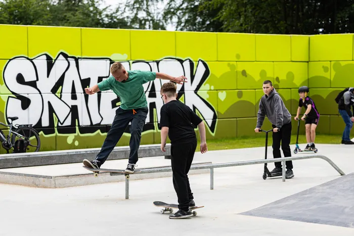 Ein Mann springt mit einem Skateboard in der Luft, während ein weiterer Skater und zwei Jugendliche auf Rollern auf dem neu eröffneten Skatepark in Halver stehen. Im Hintergrund ist ein Graffiti mit der Aufschrift 