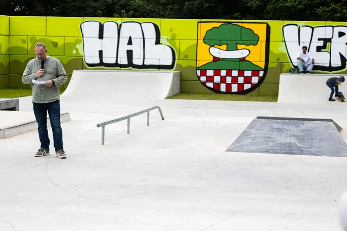 Bürgermeister Armin Kibbert hält im Halveraner Skatepark eine Rede vor einer Wand mit Graffiti und dem Stadtwappen. Im Hintergrund sind Skater auf Rampen zu sehen.