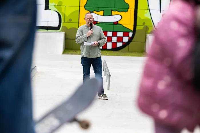 Ein Mann mit Brille hält ein Mikrofon in der Hand und spricht vor einer bunten Wand. Im Hintergrund ist ein Skatepark zu sehen.